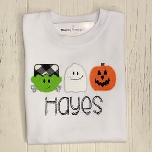 May include: White t-shirt featuring a green Frankenstein, a white ghost, and an orange pumpkin with black eyes and a mouth. The name "Hayes" is embroidered beneath the characters.