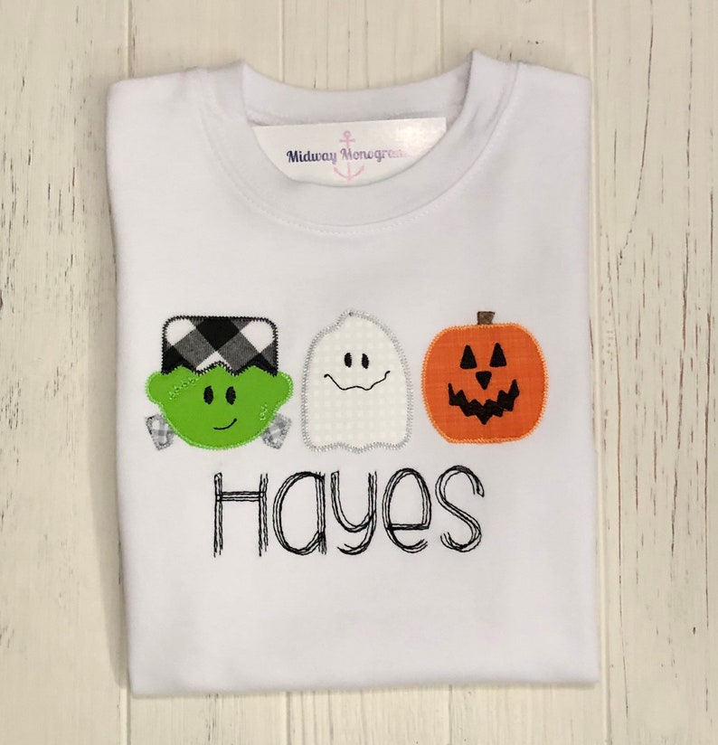 May include: White t-shirt featuring a green Frankenstein, a white ghost, and an orange pumpkin with black eyes and a mouth. The name "Hayes" is embroidered beneath the characters.