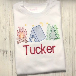 May include: White t-shirt with an embroidered design of a campfire, tent, and trees. The name "Tucker" is embroidered below the design in red.