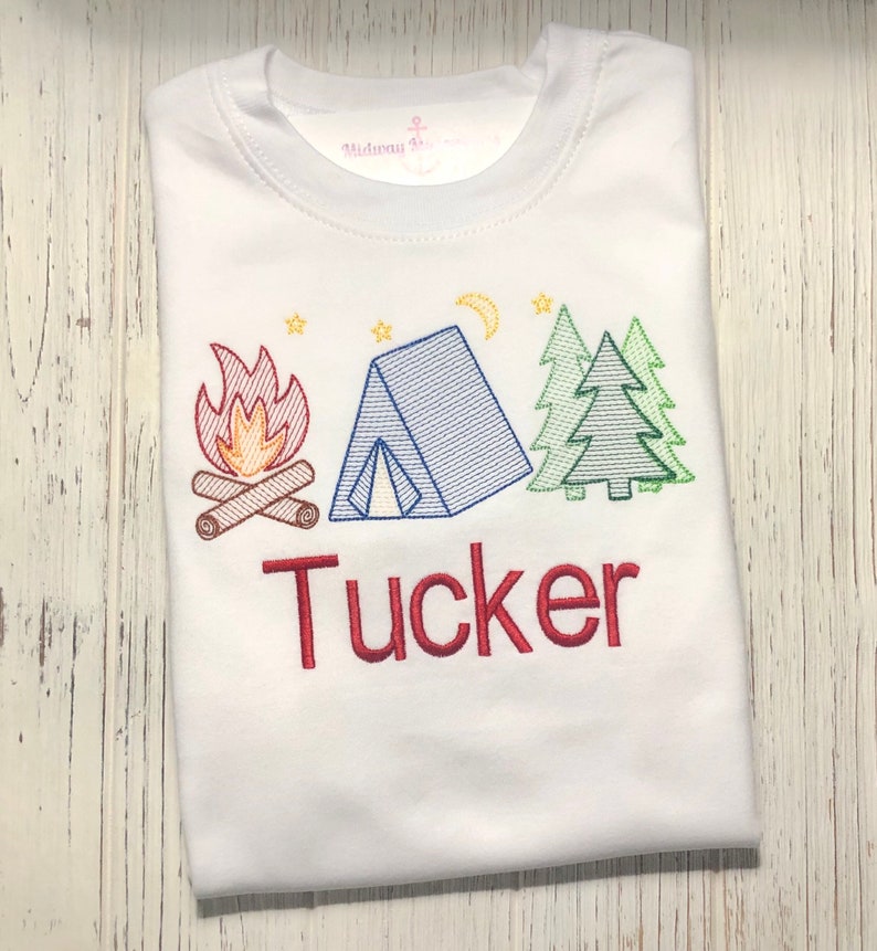 May include: White t-shirt with an embroidered design of a campfire, tent, and trees. The name "Tucker" is embroidered below the design in red.