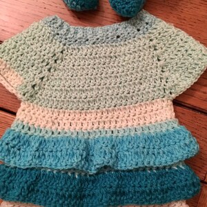 Puede incluir: Un conjunto de bebé tejido a crochet en tonos turquesa, azul y blanco. El conjunto tiene una falda con volantes y mangas cortas.