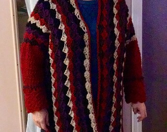 May include: A long, crocheted cardigan with a striped pattern in shades of red, purple, brown, and beige. The cardigan has a shawl collar and a loose fit.
