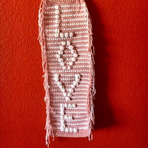 Puede incluir: Un tapiz de pared de crochet rosa y blanco con la palabra "LOVE" escrita en hilo blanco. El tapiz tiene flecos en la parte inferior y está sujeto a una varilla de madera.