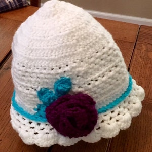 Puede incluir: Sombrero blanco de ganchillo con un adorno de flor de color morado y turquesa.