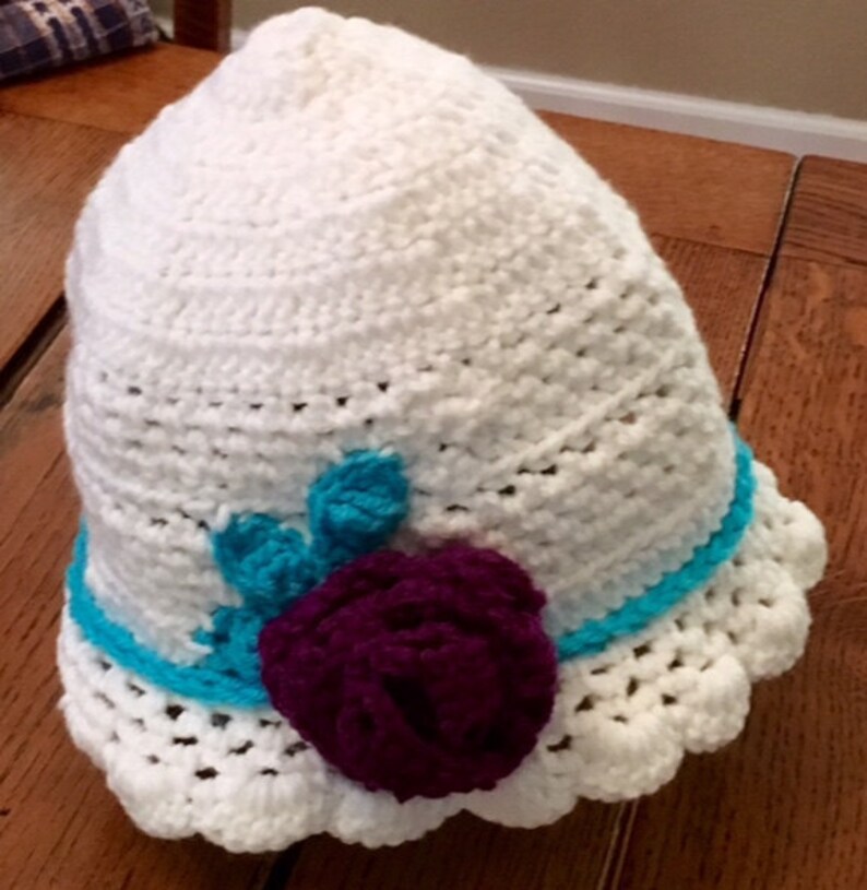 Puede incluir: Sombrero blanco de ganchillo con un adorno de flor de color morado y turquesa.