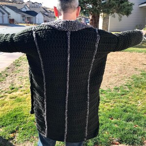 Puede incluir: Un c&aacute;rdigan largo de crochet verde oscuro con ribete gris. El c&aacute;rdigan lo lleva puesto una persona de pie frente a un barrio suburbano.
