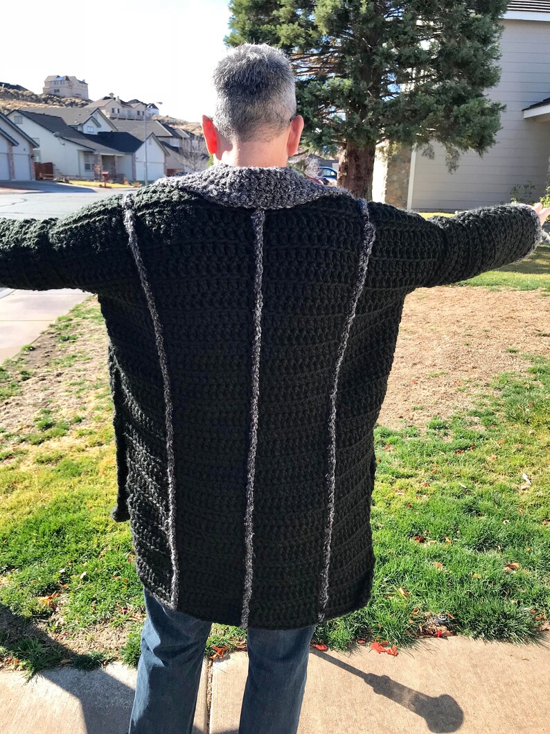 Puede incluir: Un c&aacute;rdigan largo de crochet verde oscuro con ribete gris. El c&aacute;rdigan lo lleva puesto una persona de pie frente a un barrio suburbano.
