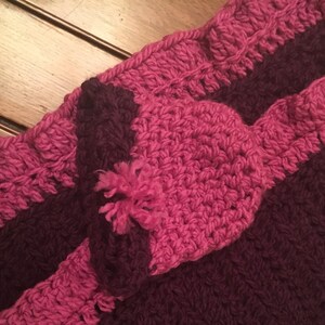 May include: A crocheted blanket with a pink and burgundy striped pattern. The blanket has a ruffled edge and a pink crocheted flower in the center.