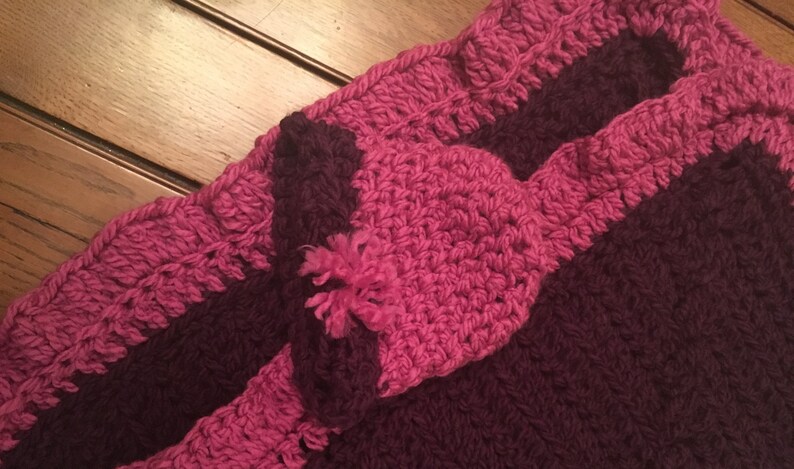 May include: A crocheted blanket with a pink and burgundy striped pattern. The blanket has a ruffled edge and a pink crocheted flower in the center.