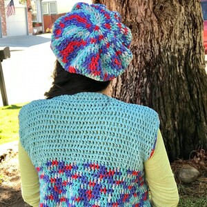 May include: A blue, red, and white crocheted beret with a swirl pattern. The beret is worn by a person wearing a blue and white crocheted vest.