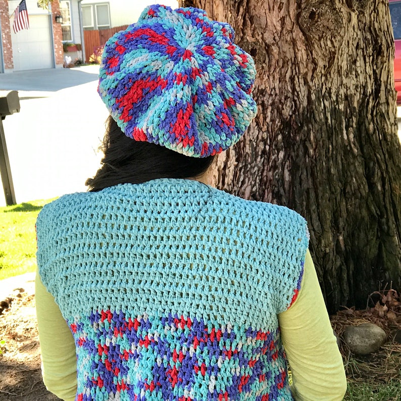 May include: A blue, red, and white crocheted beret with a swirl pattern. The beret is worn by a person wearing a blue and white crocheted vest.