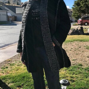 May include: A man wearing a black knit cardigan with a gray and black pattern, a black t-shirt with the text "NEW MERICA SURF BR", blue jeans, and blue sunglasses.