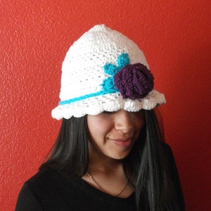 Puede incluir: Un gorro blanco de ganchillo con una flor morada y un borde turquesa. El gorro lo lleva una persona con el pelo largo y oscuro.