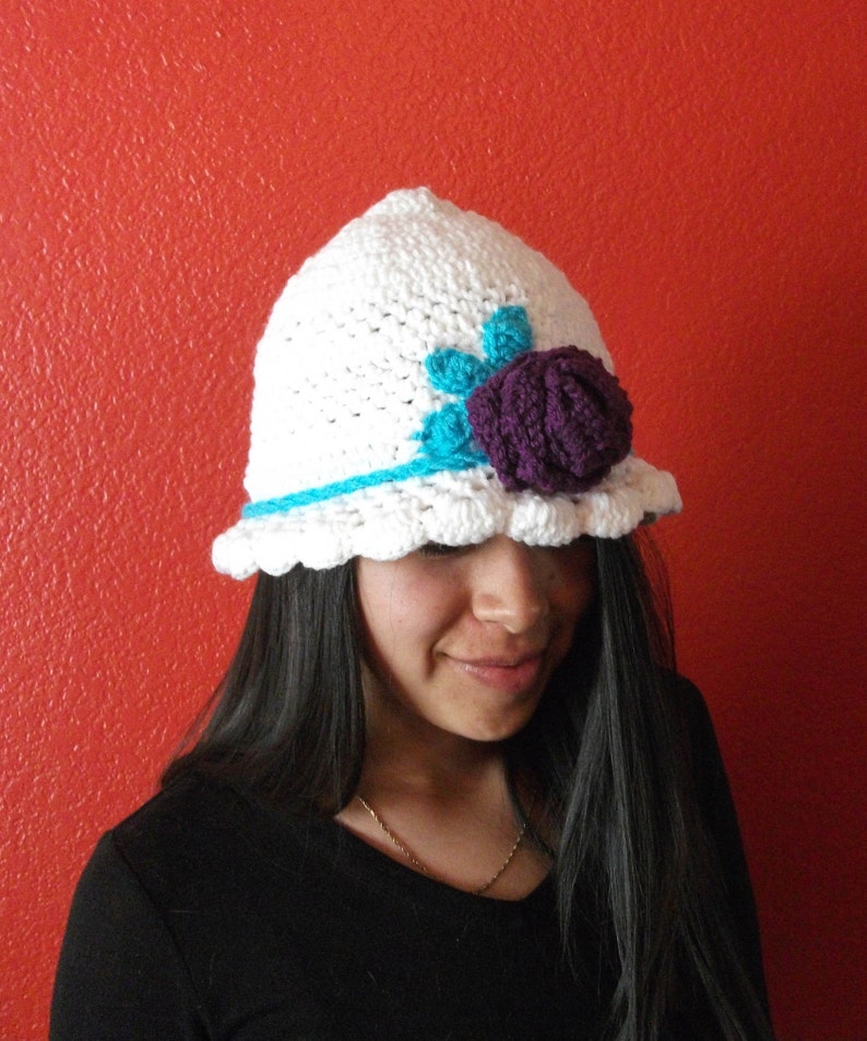 Puede incluir: Un gorro blanco de ganchillo con una flor morada y un borde turquesa. El gorro lo lleva una persona con el pelo largo y oscuro.