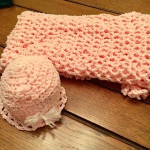 May include: A pink crocheted baby hat and blanket set. The hat has a white bow and the blanket has a delicate open stitch pattern.