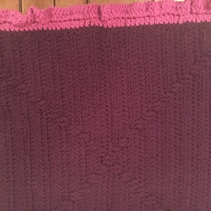 May include: A crocheted blanket with a pink border. The blanket is made with a textured stitch and is a deep purple color.
