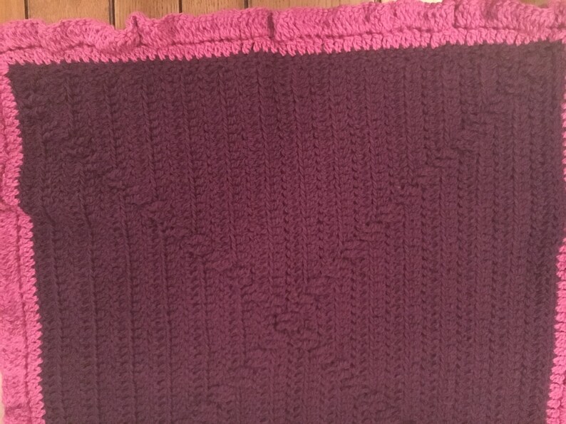 May include: A crocheted blanket with a pink border. The blanket is made with a textured stitch and is a deep purple color.
