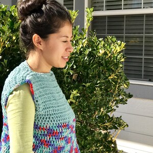 May include: A woman wearing a light blue crochet vest with a colorful geometric pattern. The vest has a scoop neck and sleeveless design.