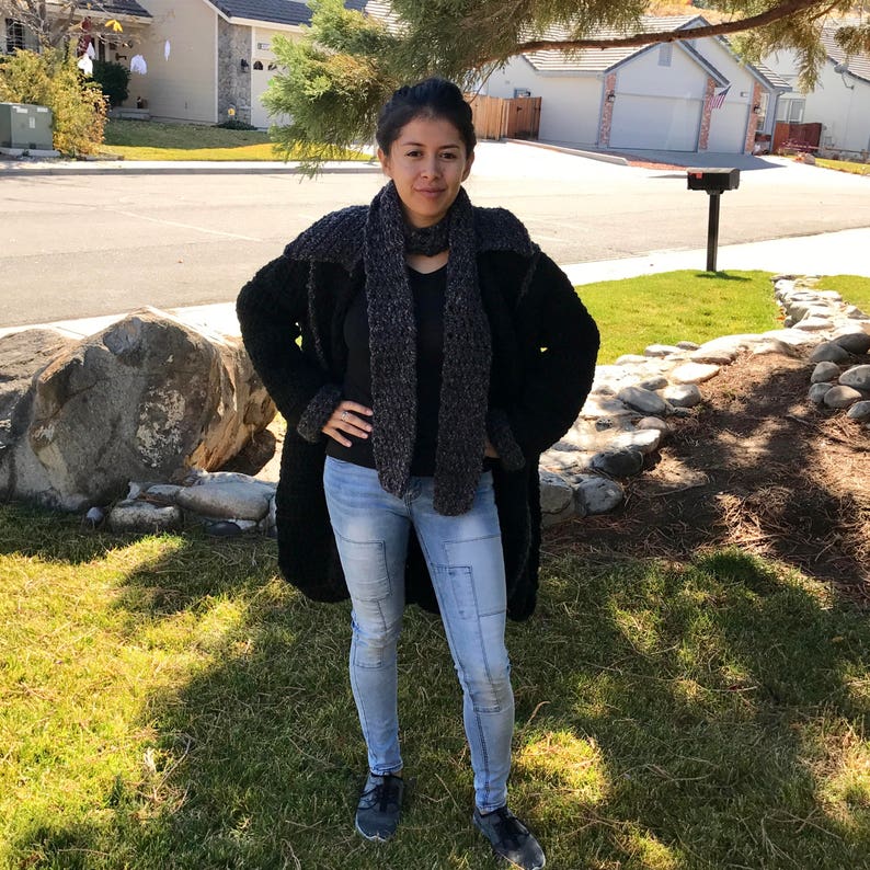 Puede incluir: Una mujer con un c&aacute;rdigan de ganchillo negro con una bufanda gris y negra, pantalones vaqueros azules y zapatillas negras.