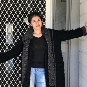 Puede incluir: Una mujer con un c&aacute;rdigan de punto negro y gris con una bufanda larga. Est&aacute; de pie frente a una puerta con una pantalla de metal negro.
