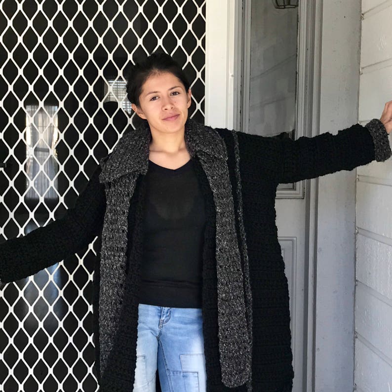 Puede incluir: Una mujer con un c&aacute;rdigan de punto negro y gris con una bufanda larga. Est&aacute; de pie frente a una puerta con una pantalla de metal negro.