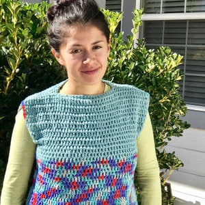May include: A woman wearing a light blue crochet vest with a colorful geometric pattern at the bottom. The vest has a scoop neck and sleeveless design.