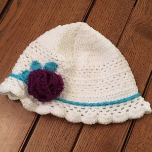 Puede incluir: Sombrero de sol blanco de ganchillo con una flor morada y un borde turquesa.