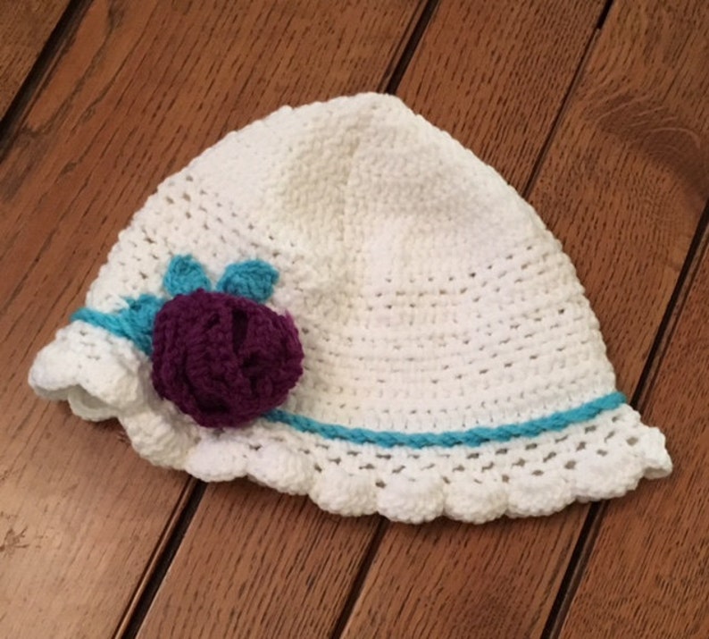 Puede incluir: Sombrero de sol blanco de ganchillo con una flor morada y un borde turquesa.