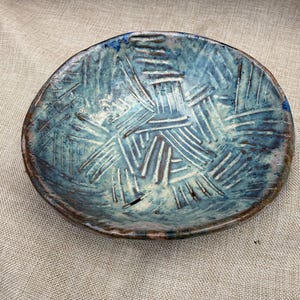 May include: A handmade ceramic bowl with a textured surface and a blue and brown glaze. The bowl has an oval shape and a unique, cross-hatched pattern. The rim is a darker brown color.