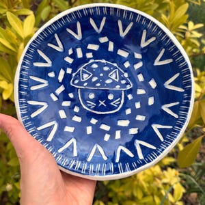 May include: A handmade ceramic plate with a whimsical mushroom cat design. The plate features a deep blue glaze with white accents, including a border of V-shaped patterns and a dotted background. The central mushroom cat illustration adds a playful touch.