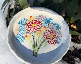 May include: A white ceramic bowl with a hand-painted floral design. The bowl features a bouquet of blue, red, yellow, and green flowers.