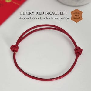 May include: A thin red leather cord bracelet featuring two adjustable knots. The text above the bracelet reads LUCKY RED BRACELET Protection · Luck · Prosperity, with a badge stating GENUINE 100% LEATHER. The design is simple and circular.