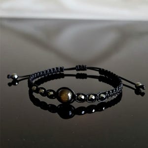 Peut inclure: Bracelet tressé noir avec une pierre centrale ronde et marron, entourée de petites perles sombres. Le bracelet a des cordons noirs réglables et des accents métalliques. Le design est minimaliste et élégant, adapté au quotidien.