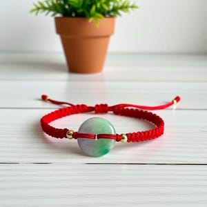 Peut inclure: Bracelet tressé rouge avec une pièce centrale en jade vert et blanc de forme ronde. Le bracelet comporte des accents dorés et des nœuds coulissants réglables. Un petit pot de plante est en arrière-plan.