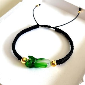 Peut inclure: Bracelet en cordon tressé noir avec un charm en forme de bok choy vert et blanc. Le bracelet comprend des perles dorées et une fermeture coulissante réglable.