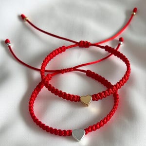 May include: Two red macrame bracelets with heart-shaped charms. One bracelet features a gold heart, the other a silver heart. The bracelets are on a white background. The bracelets are adjustable.