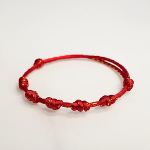 Peut inclure: Bracelet rouge avec plusieurs nœuds décoratifs. Le bracelet est fait d'un cordon rouge avec des accents dorés. Les nœuds sont espacés uniformément autour du bracelet. Le bracelet est sur fond blanc.