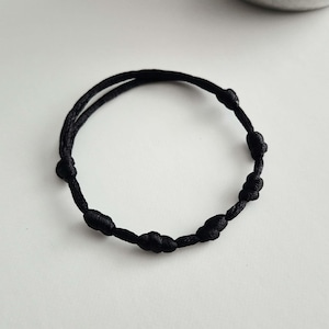 Puede incluir: Una pulsera de cordón negro que presenta una serie de nudos hechos a mano, espaciados uniformemente a lo largo de su longitud. La pulsera está construida con una sola pieza continua de material tejido fino y oscuro, formando una forma circular.