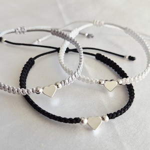 May include: Three friendship bracelets with heart charms. One bracelet is black, one is gray, and one is white. Each bracelet has a silver heart charm and silver beads. The bracelets are on a white surface.