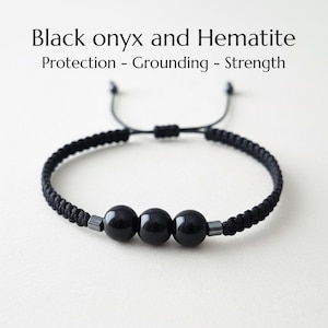 Peut inclure: Bracelet en onyx noir et hématite avec trois perles polies en onyx noir. Le bracelet est composé d'un cordon noir tressé avec des nœuds coulissants réglables. Le texte sur l'image indique : Onyx noir et hématite, Protection - Ancrage - Force.