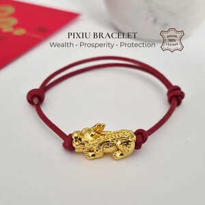 Op de afbeelding: Een rode leren koordarmband met een centrale goudkleurige Pixiu-bedel. De tekst PIXIU BRACELET Wealth · Prosperity · Protection is boven de armband zichtbaar, samen met een stempel met GENUINE 100% LEATHER.