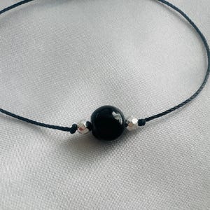 Peut inclure: Un bracelet de perles noir avec une grosse perle noire ronde et brillante et deux petites perles argentées. Le bracelet est monté sur un fin cordon foncé. Le fond est un tissu clair et argenté.