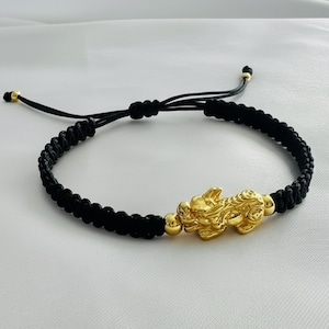 Peut inclure: Bracelet tressé noir avec un charm Pixiu doré. Le Pixiu est une créature mythique, et le bracelet a des nœuds coulissants réglables pour un ajustement personnalisé. Le bracelet est présenté sur une surface blanche.