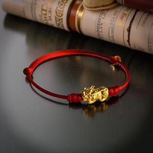 May include: A red string bracelet with a gold-colored Pixiu charm. The charm is intricately detailed and sits in the center of the bracelet. The bracelet is on a dark surface.