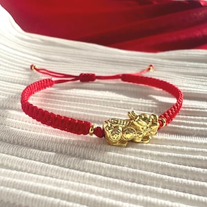 Peut inclure: Bracelet rouge tressé avec un charm Pixiu doré. Le bracelet est réglable et comporte des accents dorés. Le charm Pixiu est finement détaillé. Le bracelet est présenté sur une surface blanche texturée avec un fond en tissu rouge.