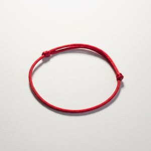 Peut inclure: Un bracelet en ficelle rouge vif, noué aux deux extrémités. Le bracelet est d'un rouge éclatant et semble être fait d'un matériau tissé résistant. Le bracelet est présenté sur un fond blanc.