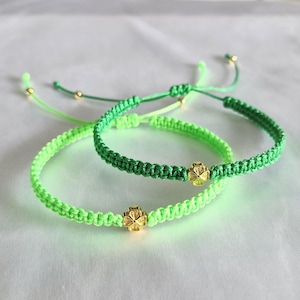 Peut inclure: Deux bracelets tressés verts avec des breloques en forme de trèfle à quatre feuilles dorées. L'un est vert vif néon, l'autre est vert plus foncé. Les bracelets ont des nœuds coulissants réglables et des perles dorées.