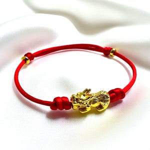 May include: A red string bracelet with a gold-coloured Pixiu charm. The bracelet has gold-coloured accents and is displayed on a white, silky surface. The Pixiu charm is a mythical creature.