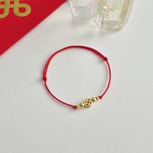 May include: A red string bracelet with gold-colored beads and a central coin-shaped charm. The bracelet is displayed on a white surface, with a red envelope and crystal-like objects in the background.