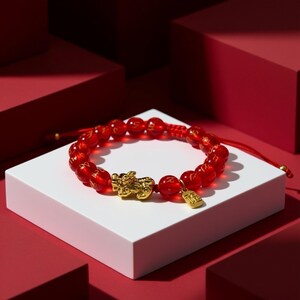Pode incluir: Pulseira de contas vermelhas com um berloque dourado com uma criatura mítica e uma pequena etiqueta retangular. A pulseira é exibida em uma superfície branca sobre um fundo vermelho.